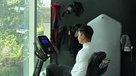 a man in athletic wear uses a recumbent stationary bike near large windows in a well-lit gym, with natural daylight and steady camera angle
