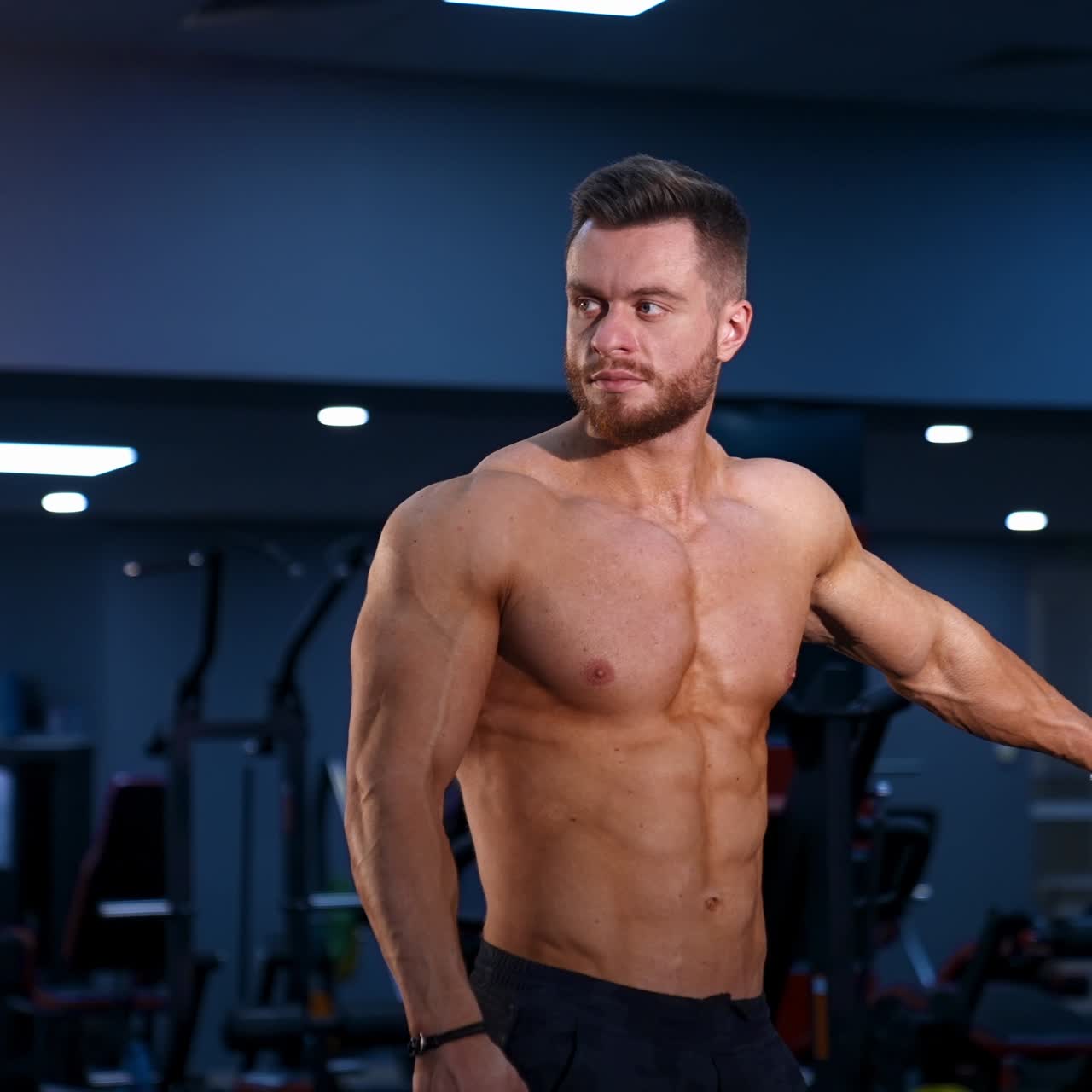 Bodybuilder is posing. Beautiful sporty guy male power. Fitness muscled man without shirt on dark gym background. Slow motion