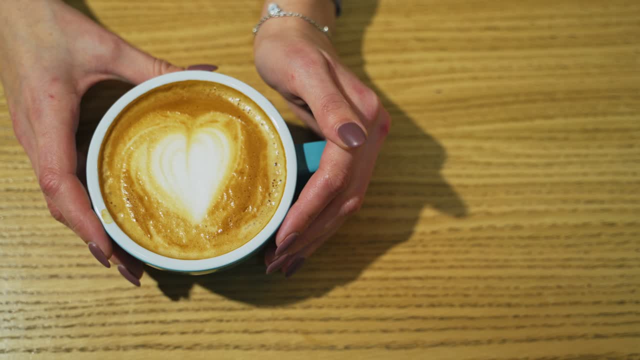 Cup with coffee on wooden background. Latte art of heart design on coffee. Female hands taking cup of hot drink from the wooden surface. Top view.