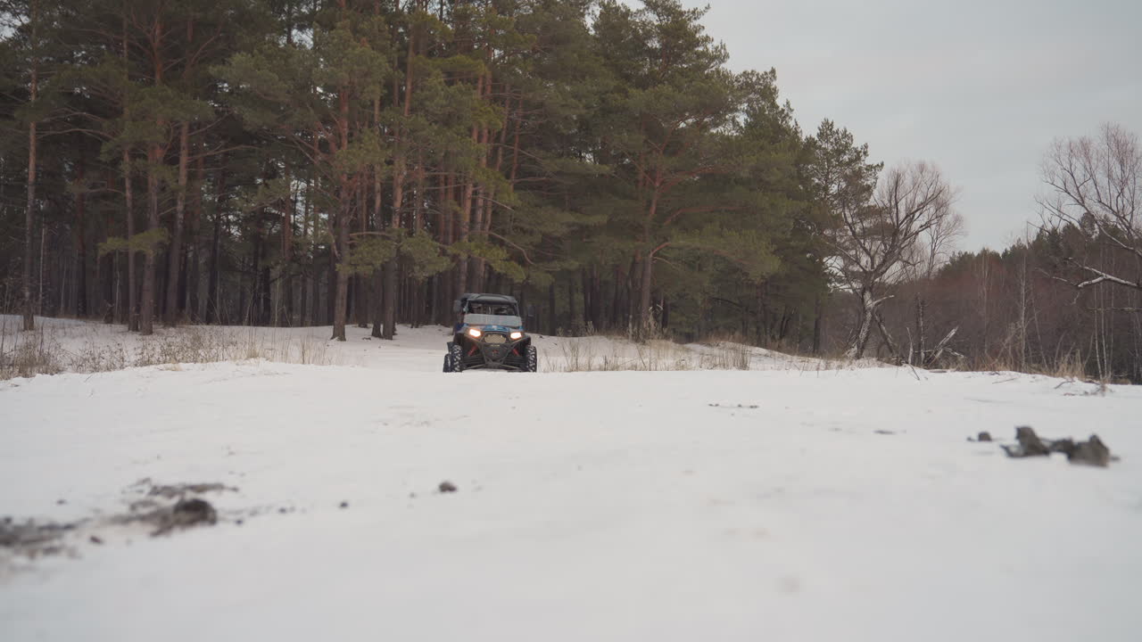 ATV che si lancia verso la telecamera attraverso la neve, con i fari abbaglianti. Prospettiva frontale, spruzzo dei pneumatici che solleva polvere, rombo intenso del motore. Il pilota che mantiene il controllo, energia da giro finale. Sfondo di pineta.