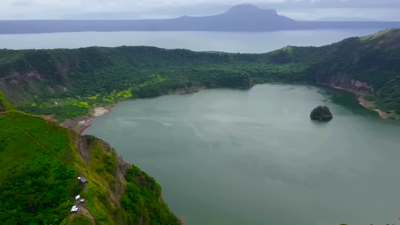 Aerial drone footage panning over Taal or Bombon lake during a cloudy day in the Philippines. The lake is surrounded by lush nature and mountains covered with thick jungle forest. large rock in water.