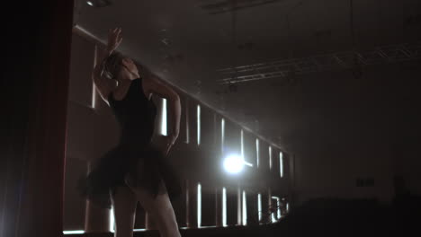 Slow-motion:-Diligent-young-graceful-ballerina-dancing-elements-of-classical-ballet-in-the-dark-with-light-and-smoke-on-the-background.-Beautiful-young-ballerina-in-darkness.