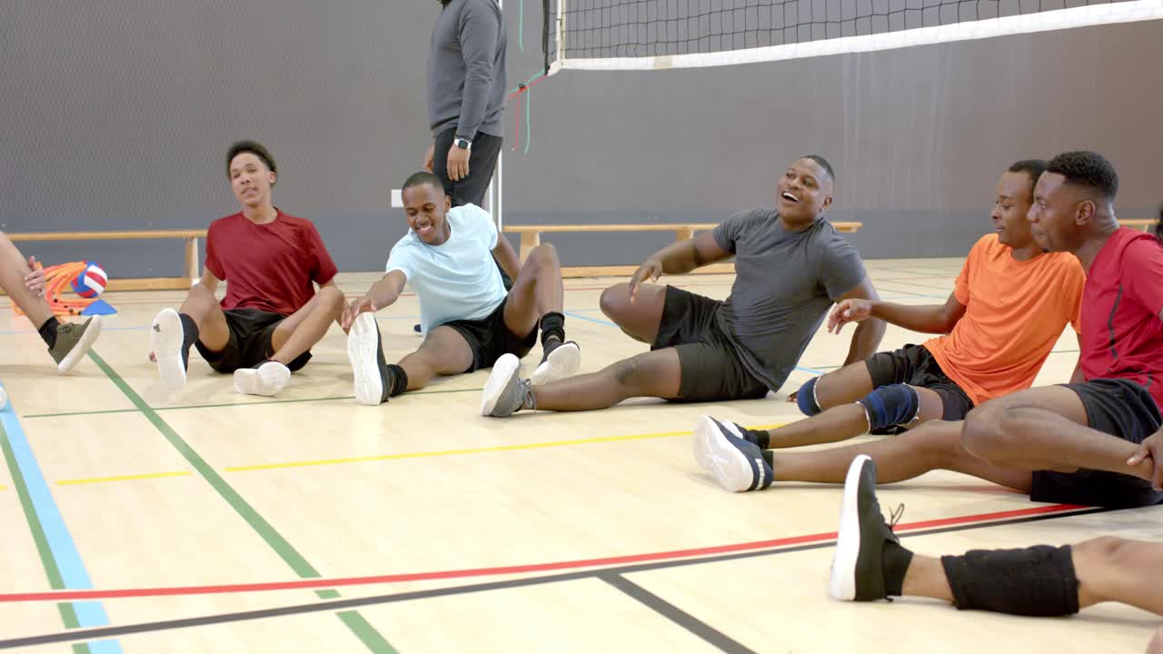 Trainer leitet Aufwärmen im Fitnessstudio an, diverses männliches Volleyballteam sitzt im Halbkreis und dehnt die Oberschenkelmuskulatur.