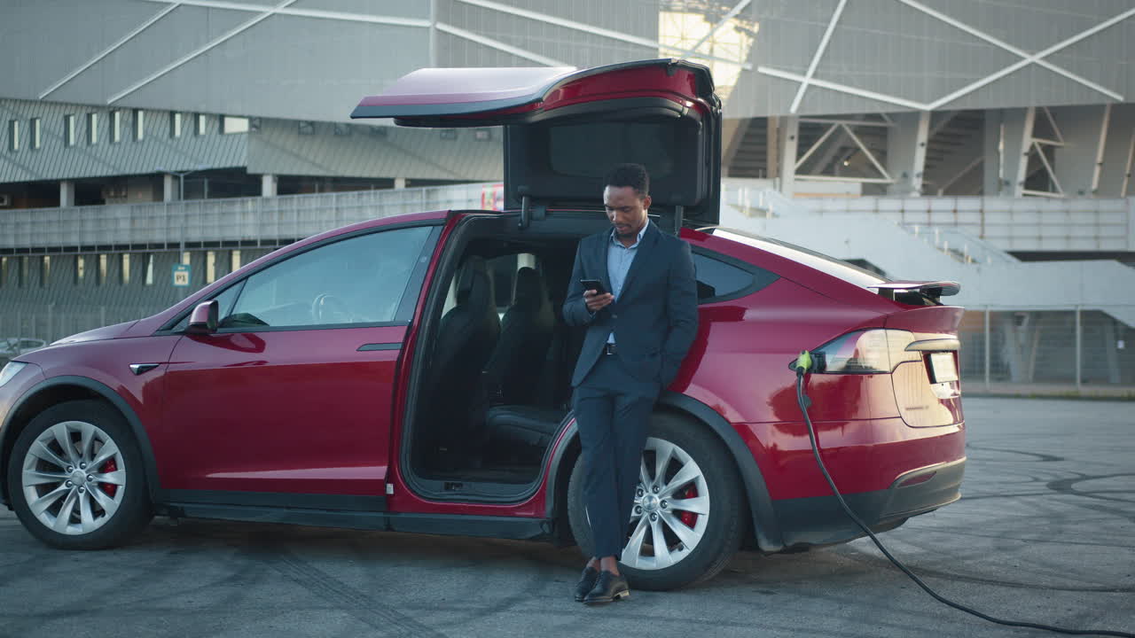 hombre de negocios usando el teléfono cerca de un vehículo eléctrico cargando
