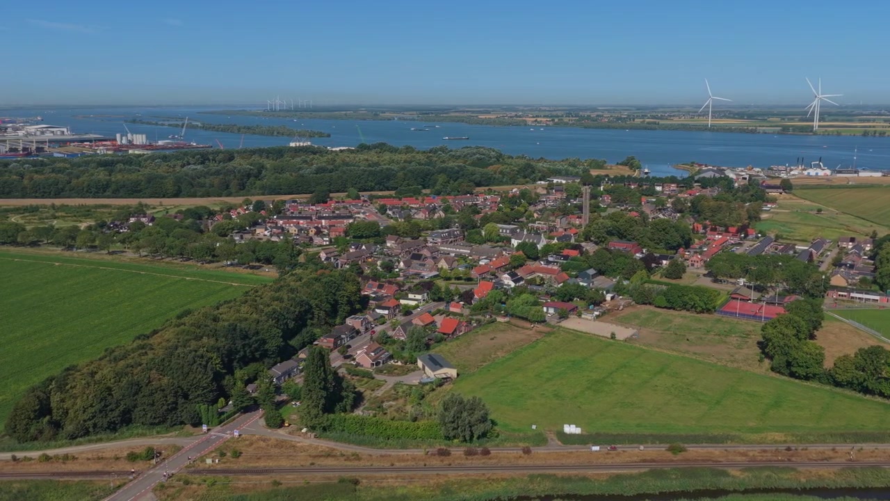 Toma aérea panorámica del pueblo holandés de Moerdijk, revelando su área residencial, campos verdes y el adyacente río Hollands Diep con turbinas eólicas