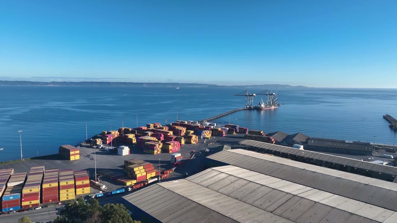 Fly-through over stacked shipping containers and a warehouse rooftop toward a distant loading pier in a calm coastal bay