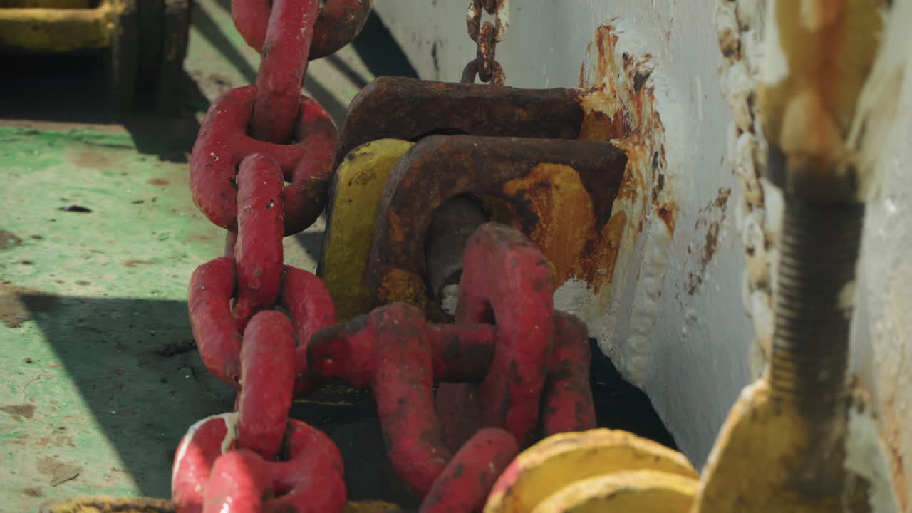 Catena rossa attraverso una staffa dello scafo arrugginita, primo piano di passacavo ossidato con vernice scrostata e macchie di sale su piastra del ponte verde, grillo pesante sotto sforzo, atmosfera portuale industriale, nautica vintage