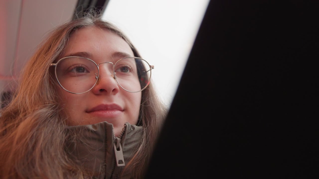 Smiling white woman on tram browsing phone and scrolling messages, wearing glasses and cozy jacket lowangle candid portrait with warm morning light, freelance creator energy and casual commuter