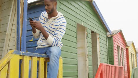 Vorderansicht-Eines-Jungen-Schwarzen-Mannes,-Der-Auf-Dem-Geländer-Einer-Hütte-Am-Strand-Sitzt-Und-Auf-Dem-Handy-SMS-Schreibt,-4k