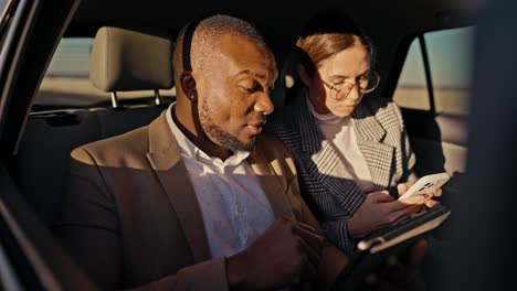 A confident man with Black skin and a beard a businessman in a brown suit explains his plans and proposals to a businesswoman in round brunette glasses using his tablet during a general trip in the cabin of a modern car
