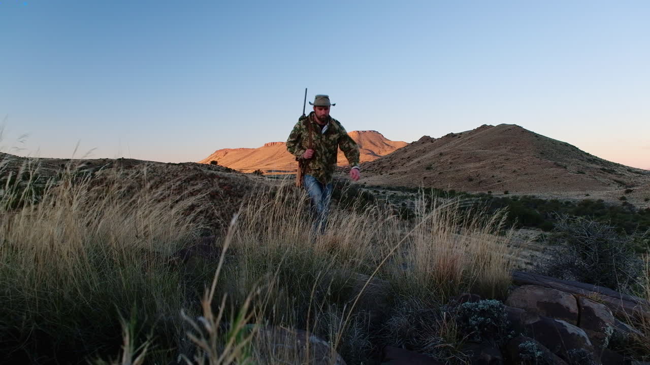 어깨 위에 소총을 들고 있는 사냥은 광활한 카루 풍경에서 바위 언덕을 고 있습니다.