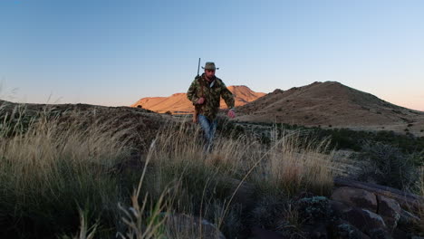 hunter with rifle over shoulder walks up rocky hill in vast karoo landscape