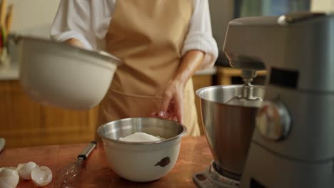 a woman baking in a kitchen, whisking flour in a bowl with a stand mixer in the background.