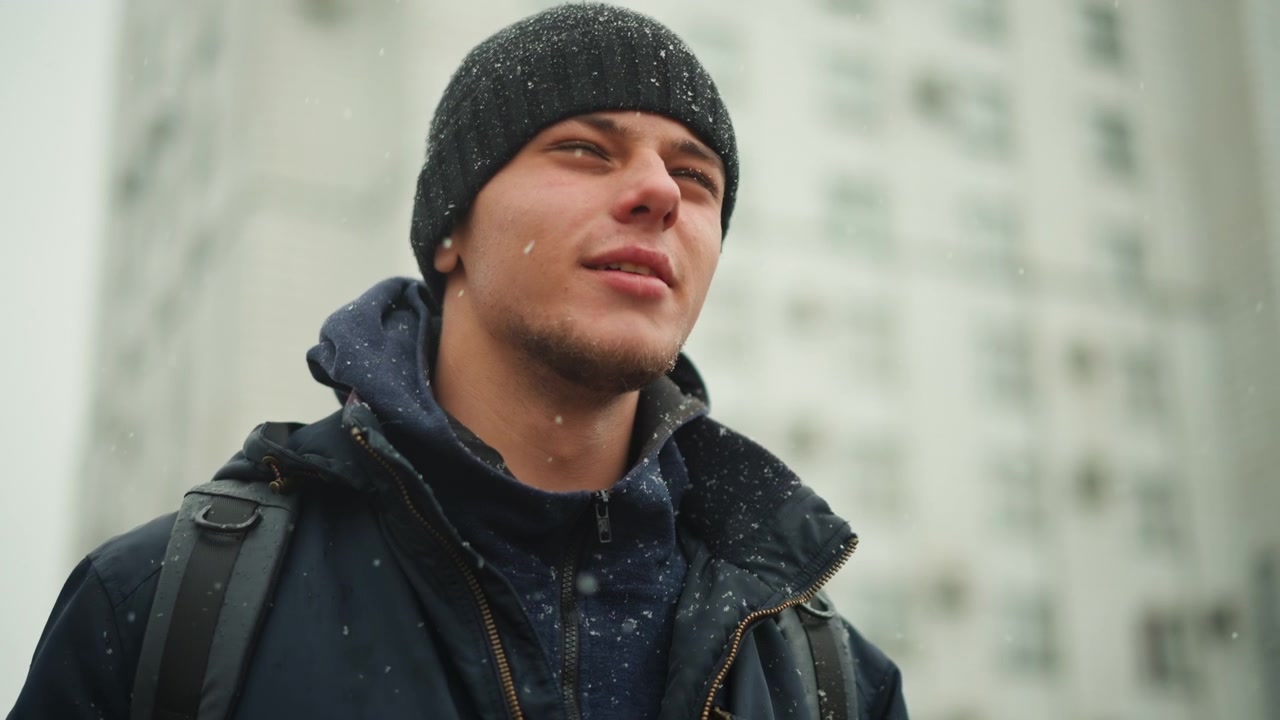 Primo piano di un uomo che indossa abiti invernali con la neve che gli cade intorno. Il soggetto è vestito con abbigliamento esterno scuro, inclusi giacca e berretto, mentre porta uno zaino. Sono visibili edifici alti.
