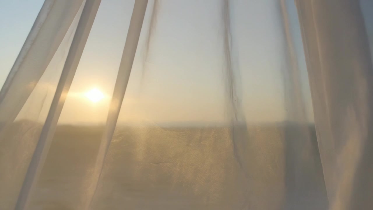 Billowing sheer curtains pushed by breeze across window in room, revealing low sun near horizon