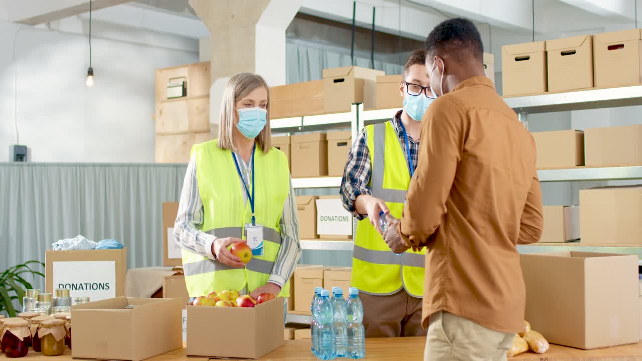 자선 창고에 있는 노숙자에게 음식과 물을 주는 안면 마스크를 쓴 백인 젊은 남성과 노인 여성 자원봉사자