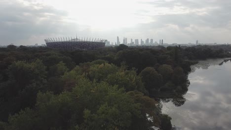 Toma-Aérea-De-Drones-Del-Horizonte-De-Varsovia-Con-Estadio-Nacional,-Bosque,-Lago-En-Un-Día-Soleado