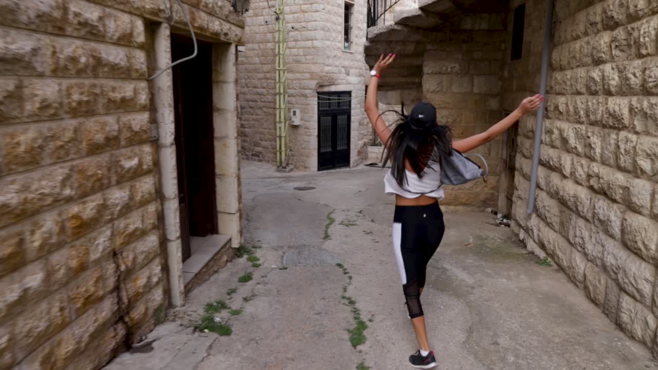 Giovane donna libanese che salta e saltella felicemente in uno stretto vicolo cittadino con muri in pietra, panoramica ravvicinata da dietro