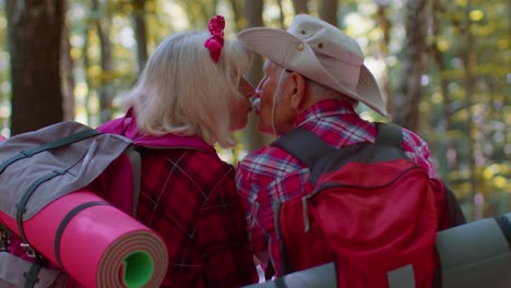 Grandmother-grandfather-senior-tourists-hikers-sitting-on-tree-and-hugging,-kissing-in-summer-wood