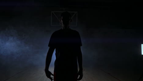 Ein-Basketballspieler-In-Sportkleidung,-Roten-Shorts-Und-Einem-Blauen-T-Shirt-Geht-Im-Gegenlicht-Auf-Einen-Dunklen-Basketballplatz,-Kommt-Aus-Dem-Rauch-Und-Wirft-Einen-Basketballball-Auf-Den-Boden,-Während-Er-In-Zeitlupe-In-Die-Kamera-Schaut