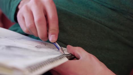 Close-up-of-artist's-hands-drawing-picture-and-holding-sketch-pad-while-sitting-on-pink-couch