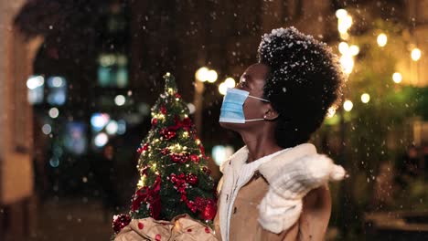 close-up view of joyful african american woman wearing facial mask holding a christmas tree and playing with the snow on the street in christmas