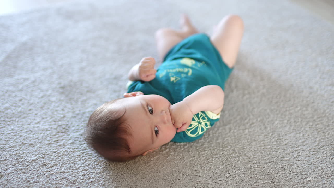 Thoughtful calm kid lies on the carpet. Baby boy wearing blue romper holding his fist in his mouth. White backdrop.