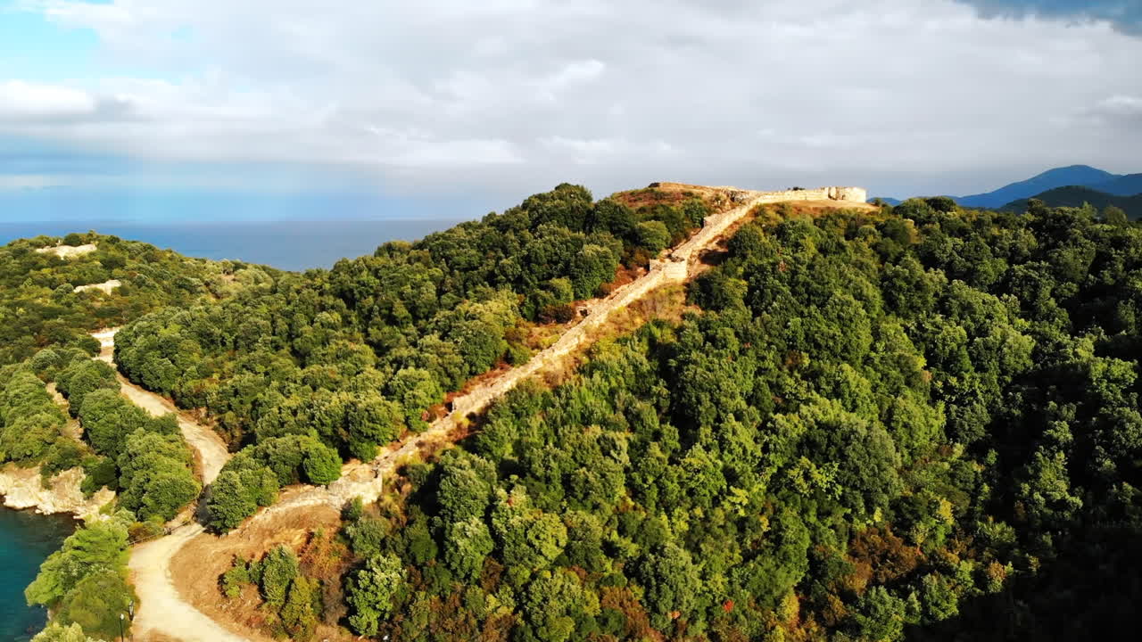 Hills covered with lush greenery. Remains of ancient structures on the top on the hill. Aegean sea coast. Road. Greece