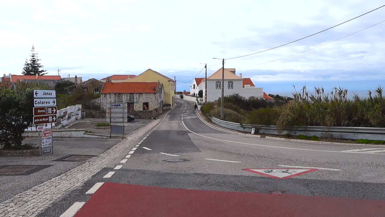 Een leeg kruispunt in een dorp vlak bij de Atlantische kust toont traditionele huizen, wegmarkeringen en gedempte tinten onder een bewolkte hemel, wat een kalme en ietwat vervallen sfeer creëert.