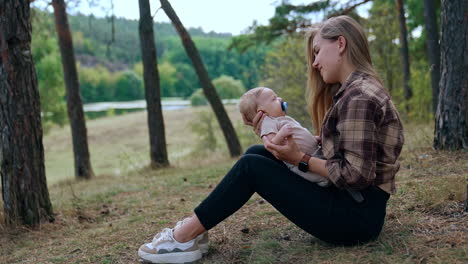 caucasian woman with blond long hair sits on the ground in the forest. mother kisses her lovely tiny baby with pacifier in mouth.