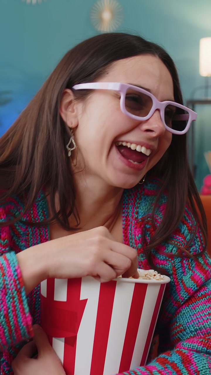 Young woman sits on sofa eating popcorn watching interesting tv serial sport game online at home