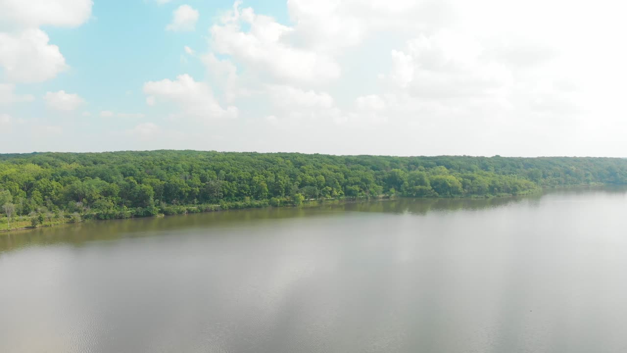 일리노이 숲 4k에서 흰색 푹신한 구름과 푸른 하늘이 있는 밝은 녹색 잎이 있는 호수 위의 수평선과 나무 줄을 향해 드론 전진 비행