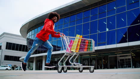 man happily running with a shopping cart full of bags outside a modern mall