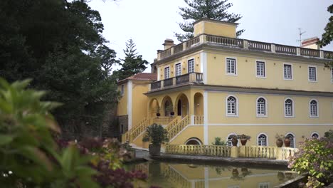 Establishing-shot-of-yellow-facade-of-Colonial-house-with-scenic-lake,-Flowers-in-the-foreground