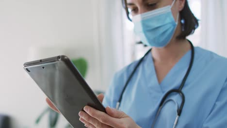 Caucasian-female-doctor-wearing-face-mask-at-home-using-tablet