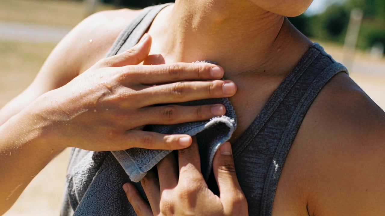 Donna con canotta sportiva grigia che si asciuga al parco, rimuovendo il sudore post-corsa con un asciugamano