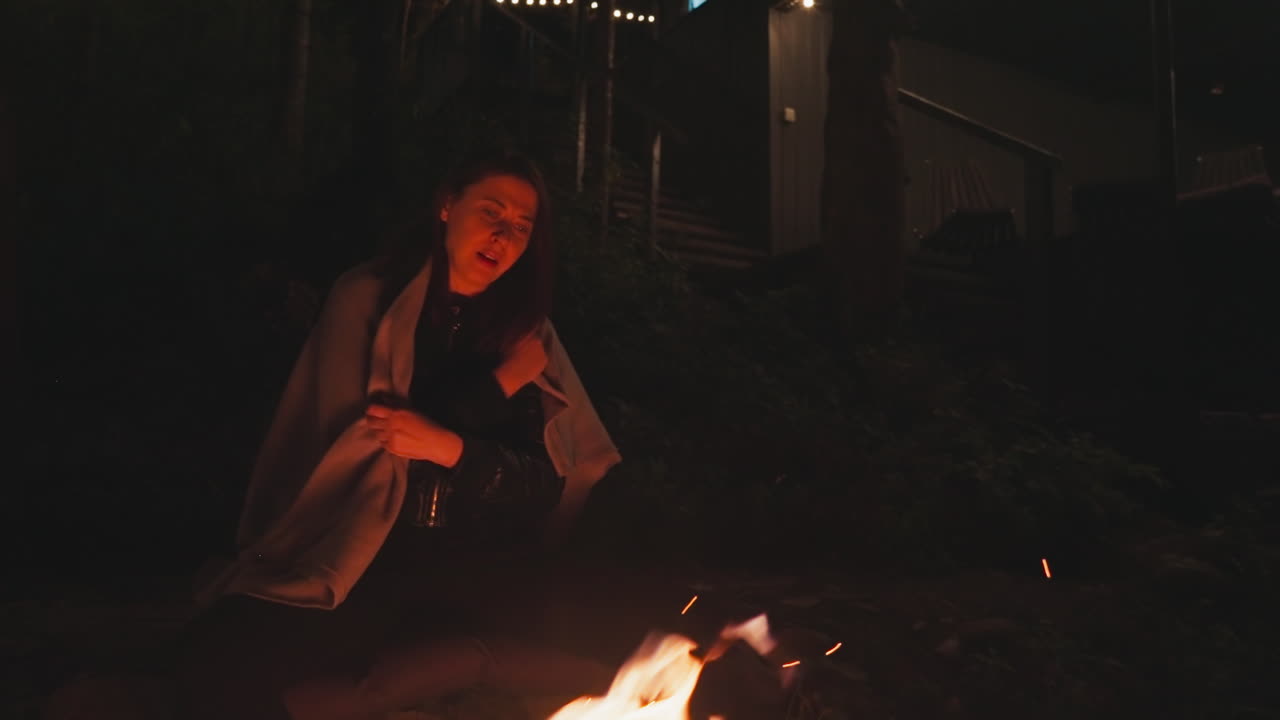 молодая женщина смотрит на огонь поздно ночью. жена настраивает одеяло на плечи, сидя на камнях у костра. время погрузиться в царство мечт и чаяний.