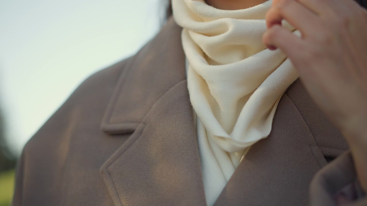 Primo piano di una donna che indossa una sciarpa color crema e un cappotto marrone, mentre si sistema la sciarpa con la mano. Una luce soffusa evidenzia la texture del tessuto su uno sfondo esterno.