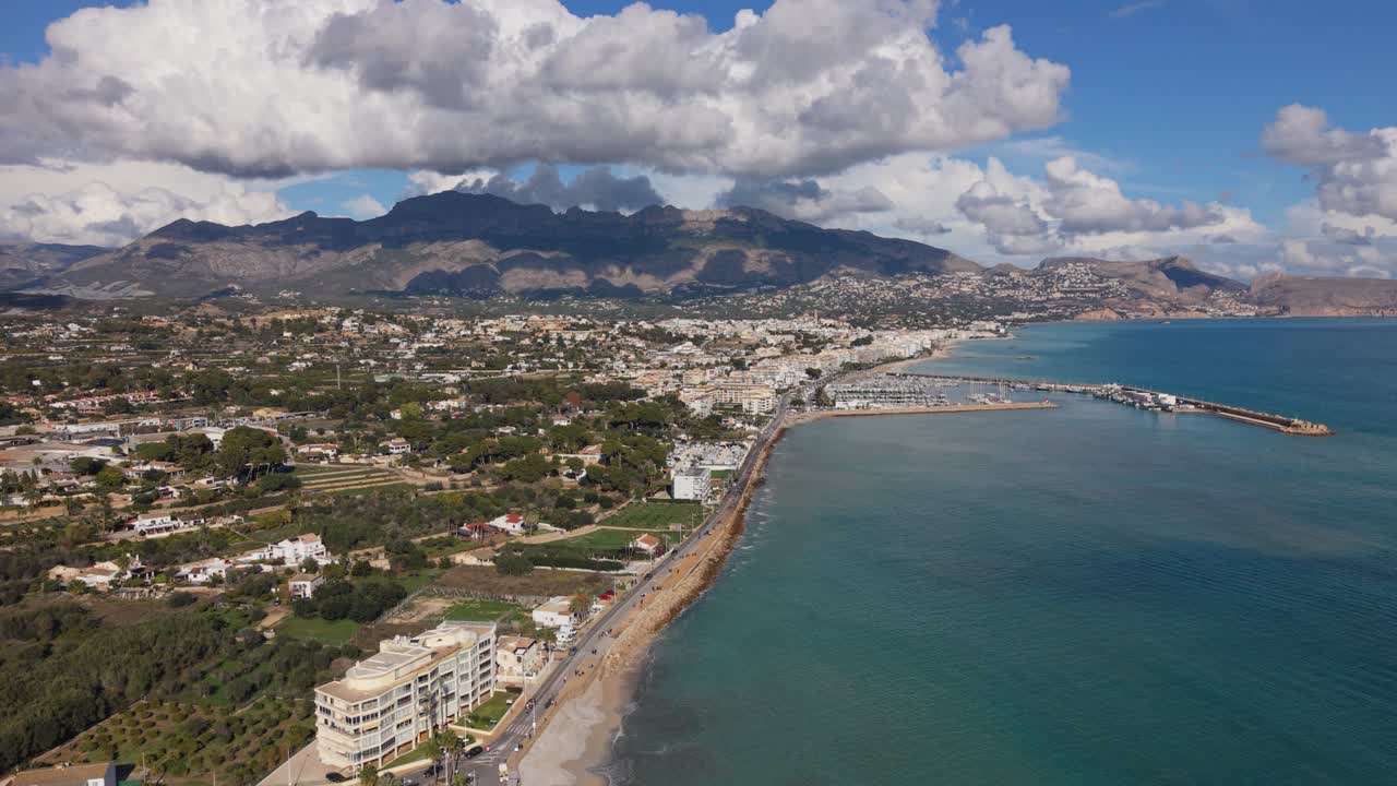 Albir e Altea, Costa Blanca, Espanha. Filmagem aérea de drone para trás da orla marítima de ambos os resorts com porto e marina sobre água azul calma e um cenário de montanhas e nuvens interessantes sob sol forte