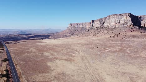 Aerial-drone-shot-descending-down-to-a-remote-rural-road-which-navigates-around-a-spectacular-sandstone-mountain-in-the-Drakensberg,-KwaZulu-Natal,-South-Africa