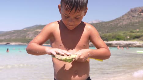 Close-up-slow-motion-on-cute-caucasian-greek-child,-filling-a-toy-shovel-with-brown-sand,-beautiful-beach-on-the-background,-summer-of-2020-at-Stoupa-beach,-Peloponnese,-Greece-