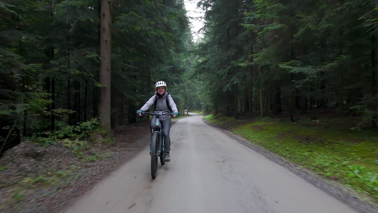 Woman rides electric mountain bike on dirt track through forest, backward dolly