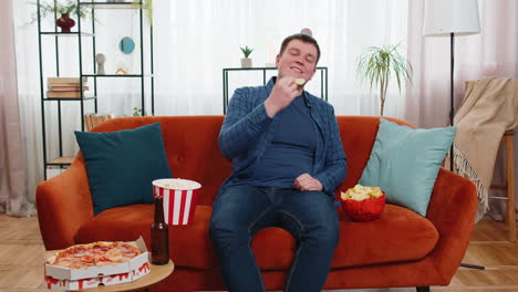 happy fat man sitting on sofa eating junk food chips pizza popcorn and beer watching tv film game