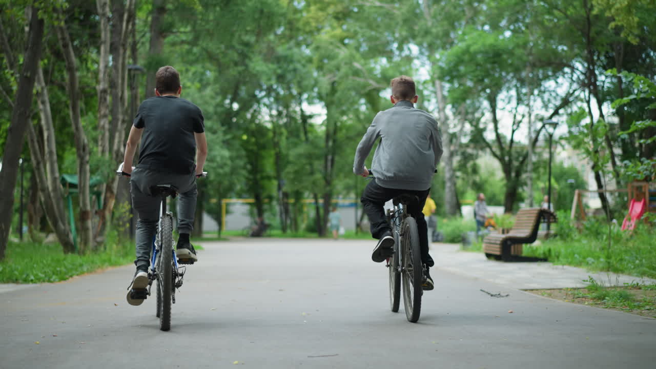 나무로 둘러싸인 푸른 공원을 가로질러 함께 자전거를 타고 다니는 두 친구의 뒷면, 두 사람은 길을 따라 동기화되어 달리고 있으며, 멀리서 걸어가는 사람들의 흐릿한 인물이 있습니다.