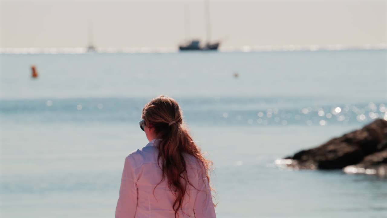 Smiling woman wearing sunglasses and a white shirt stands on the beach under the sun, with the sea glittering behind her