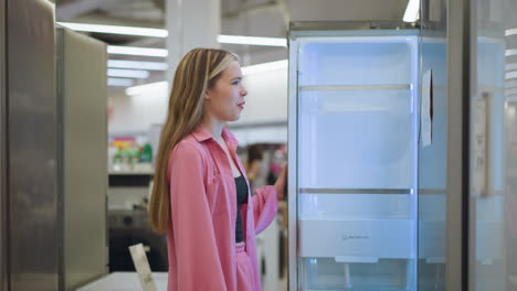 lady wearing pink dress opens refrigerator in a brightly lit store, the refrigerator interior glows with beautiful illumination, and she slides her hand on its edge, inspecting it closely