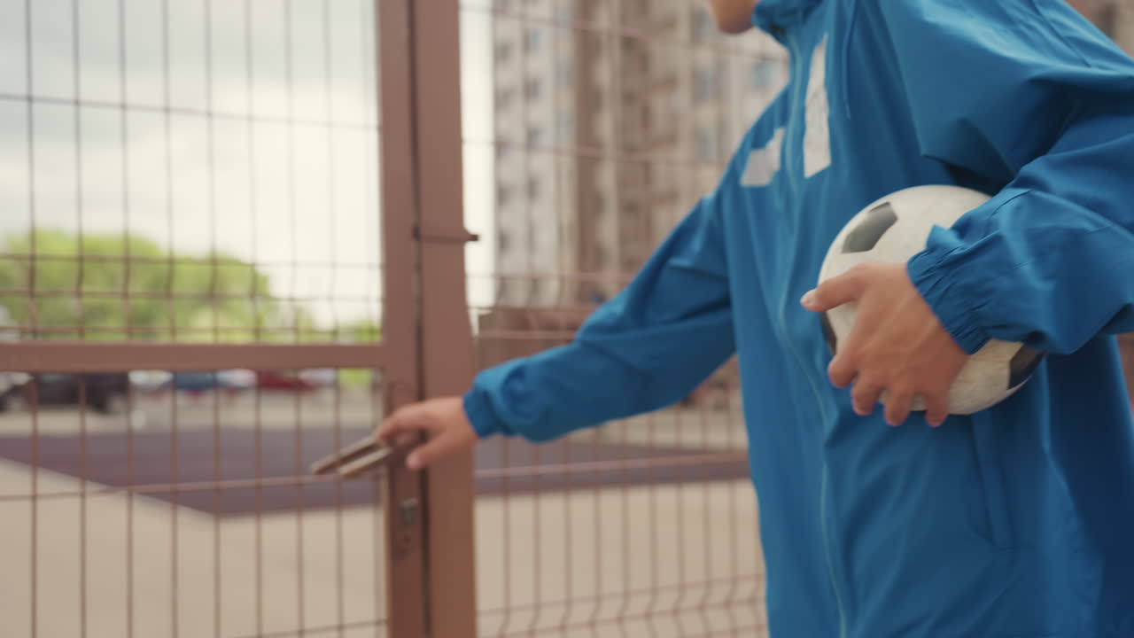 person holding soccer ball by gate with blue rain jacket, metal fence and latch, apartment buildings in background, overcast sky, quiet mood of pregame arrival and preparation for practice