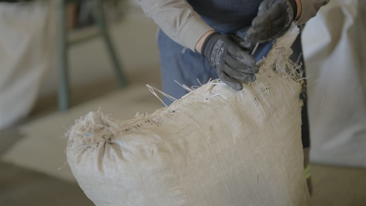 A worker in gloves seals a large, full white bag at a yerba mate factory. Close-up action.