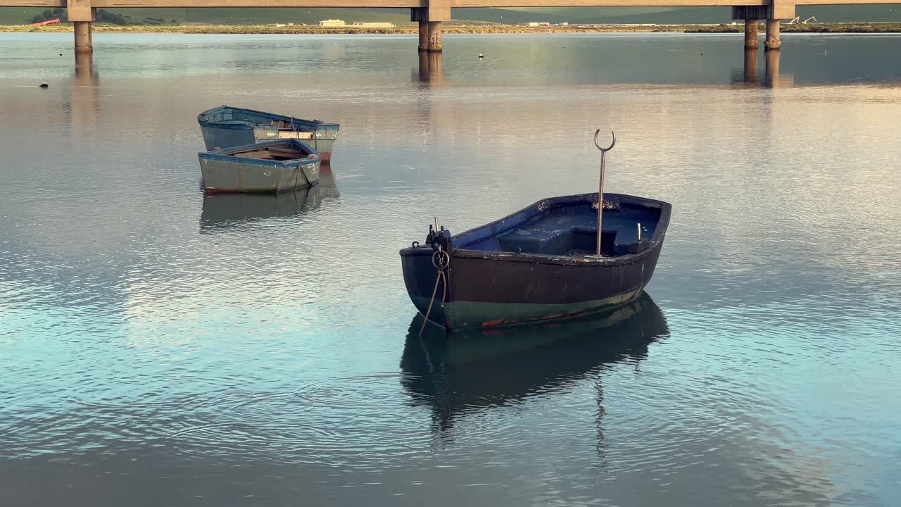 tres barcos de pesca desgastados flotan suavemente en las aguas tranquilas de barbate, andalucía, españa, reflejando el cielo sereno y el puente en el fondo, creando una escena marítima pacífica.