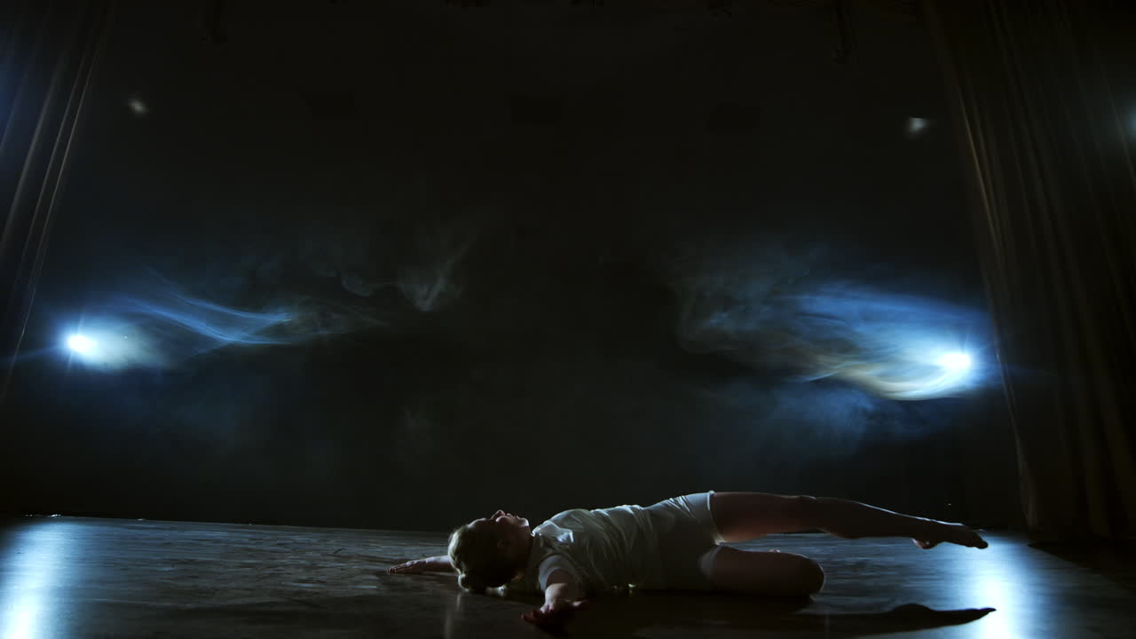 Modern dance woman in a white dress dances a modern ballet jumps makes rotation on the stage with smoke in the blue spotlights